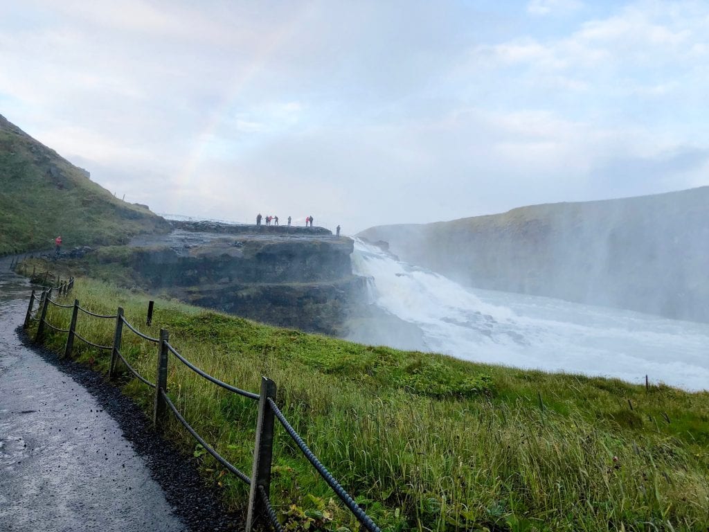 A trail leading up to a waterfall and a group of people marveling at a rainbow in the distance. 