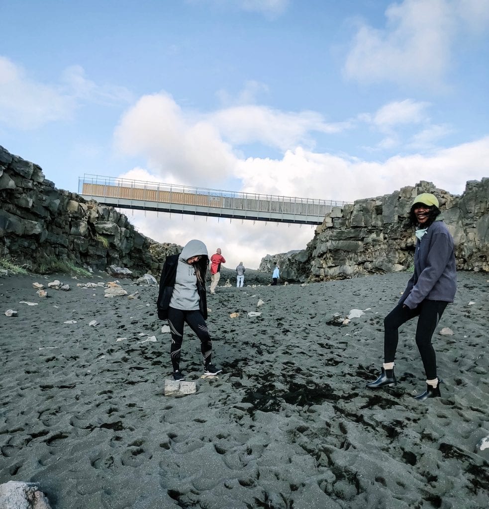 Two women standing on the black sand in front of the Bridge Between Continents in Iceland. 