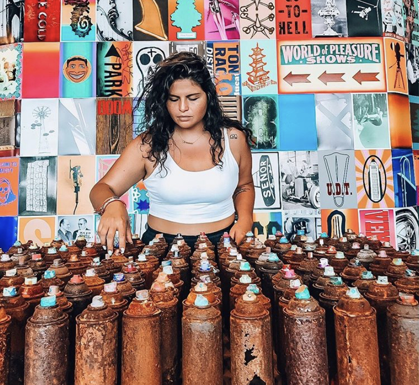 Brown woman standing in front of rusty spray paint bottles. 