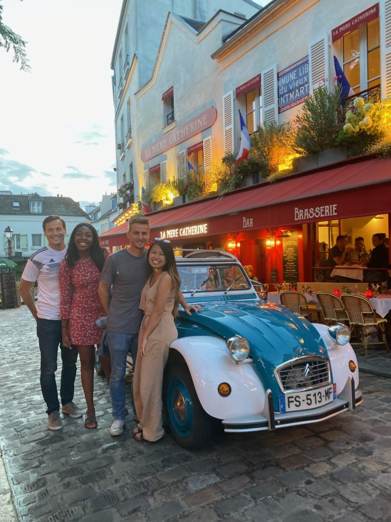A white boy, black girl, white boy, and asian woman standing in front of a blue car in Montmartre Paris.