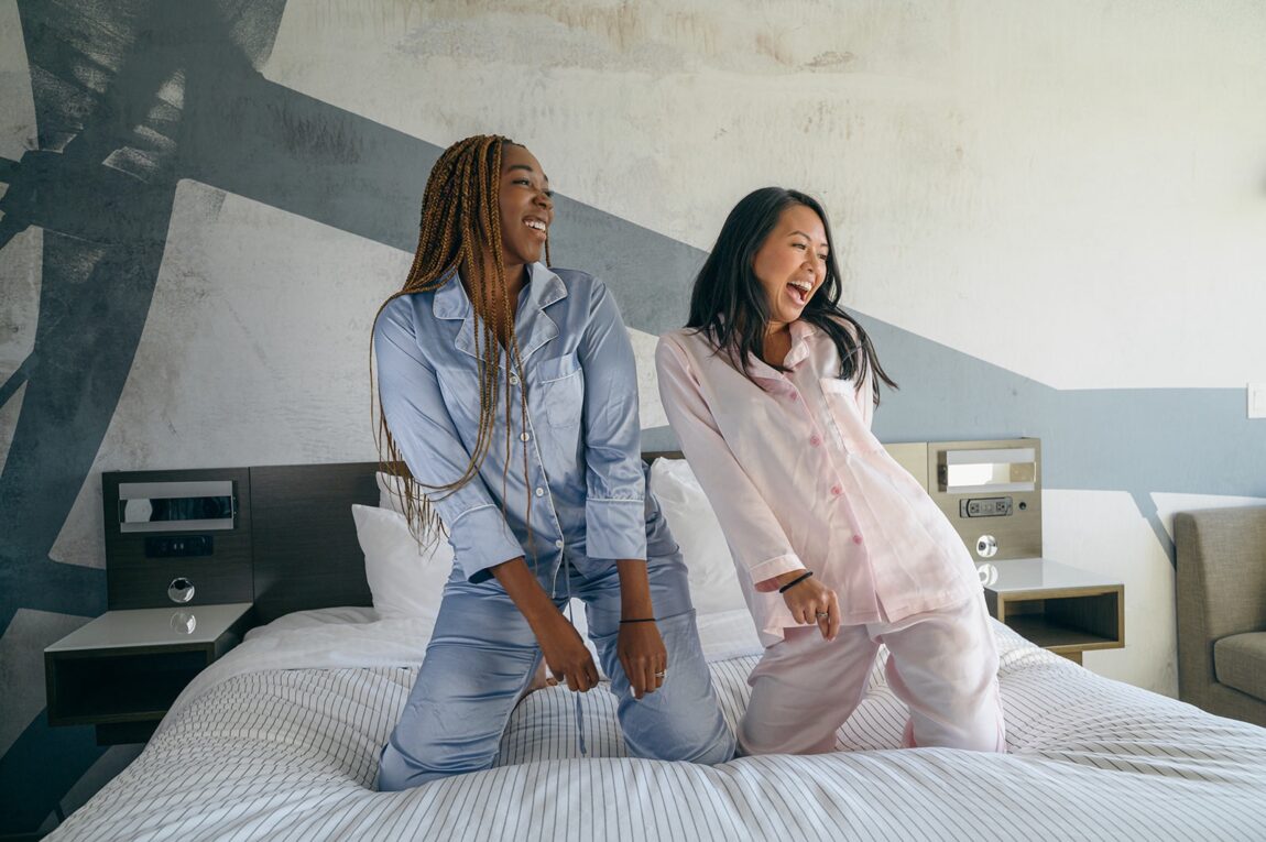 Photo of two young women kneeling on a bed in a hotel looking to the left