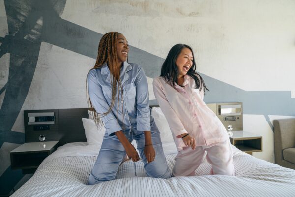 Photo of two young women kneeling on a bed in a hotel looking to the left